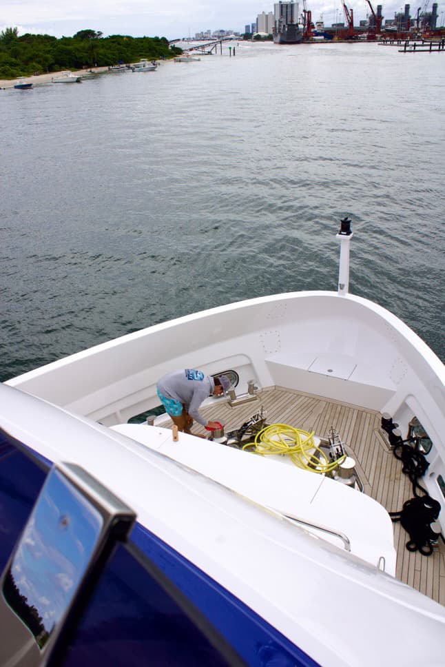 Crew member tending the foredeck of a white motor yacht on a waterway with boats and industrial cranes ahead, Miami area.