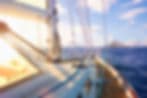 Sailboat deck with rigging and sails overlooking open blue water and distant islands.