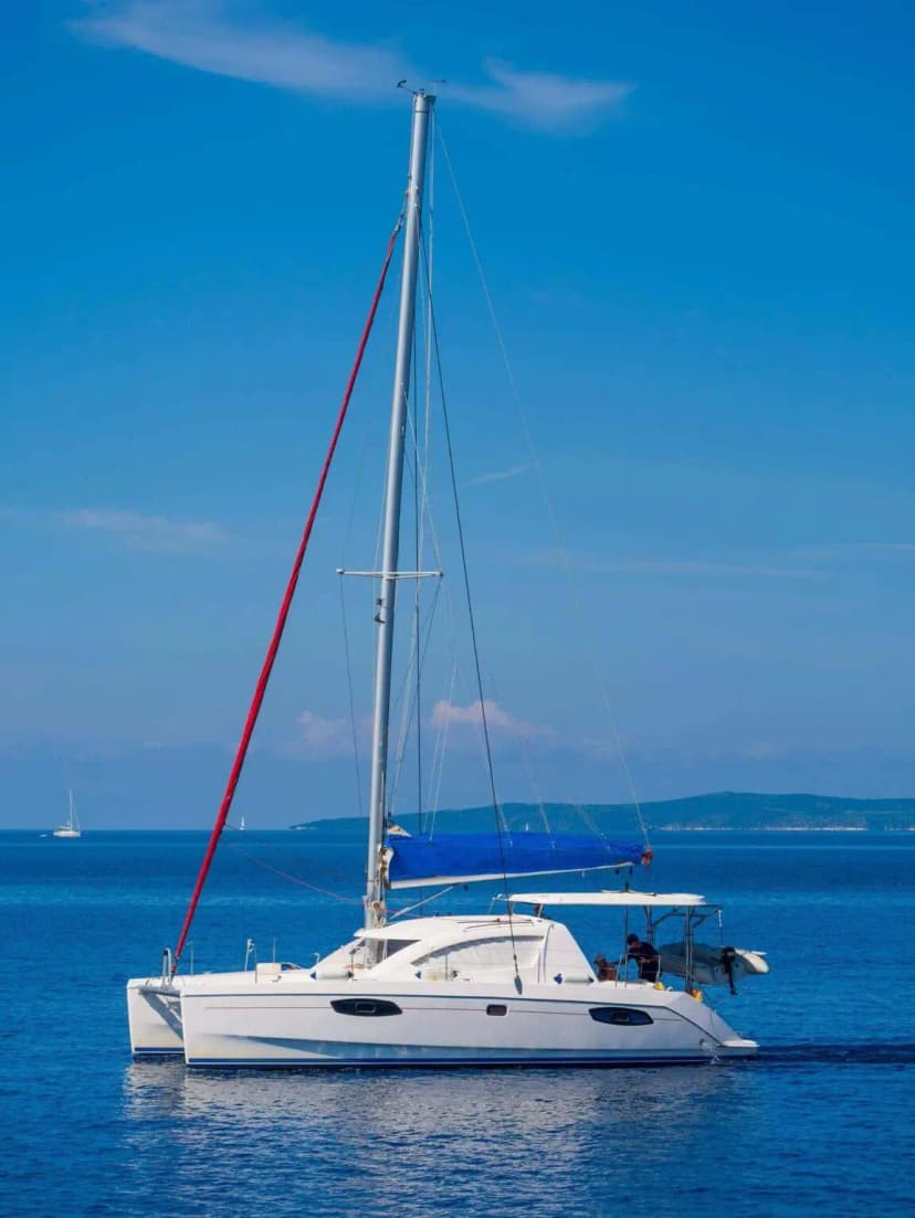 White sailing catamaran with a blue canopy and red rigging anchored on calm blue water, with a distant island on the horizon.