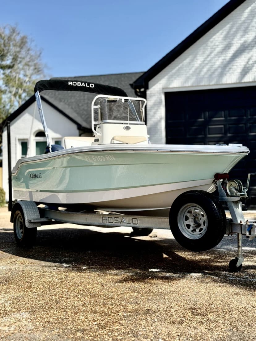 Light green Robalo center console boat on its Robalo trailer parked in a driveway beside a white brick home.