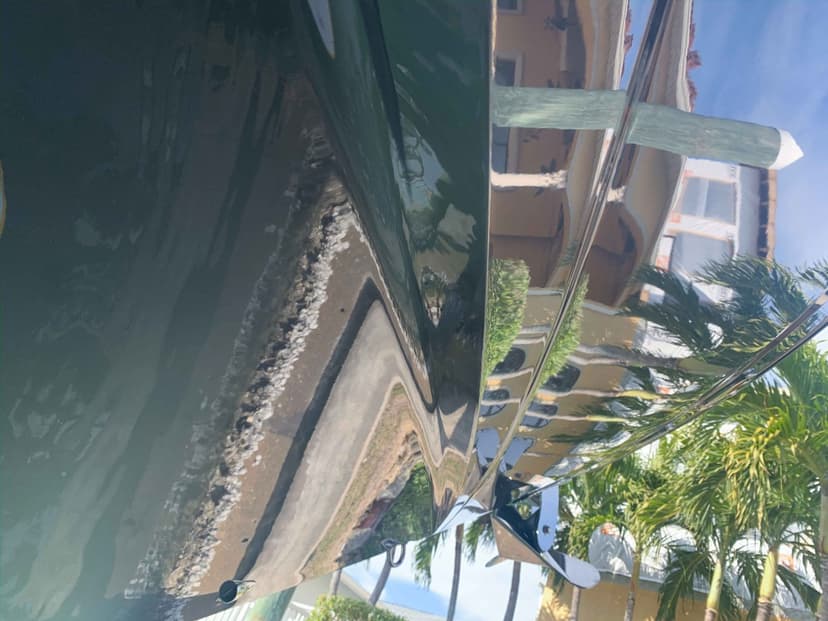 Polished green boat hull and stainless steel railing with reflections of a canal, palm trees, and waterfront homes.