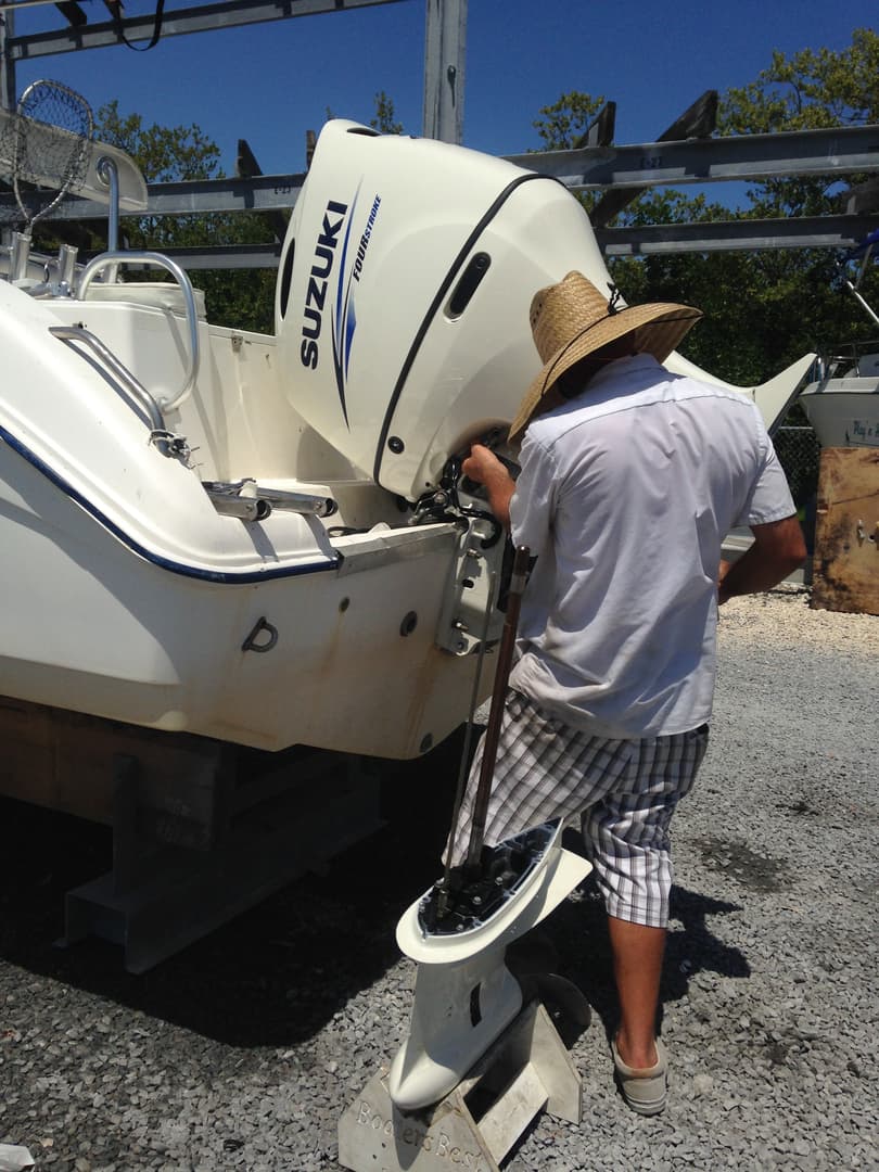 A technician in a straw hat working on a white Suzuki four-stroke outboard motor on a boat at a gravel boatyard, with the boat on a trailer stand.
