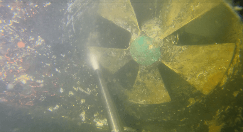 Underwater photograph of a five-blade bronze propeller being cleaned with a water-jet wand, marine growth visible on the blades.